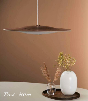 Een minimalistische hanglamp met een dunne, sculpturale houten kap hangt boven een ronde tafel. Op de tafel staat een houten dienblad met een witte keramische vaas met delicate gele bloemen en een kleine glazen vaas met gedroogde grassen. De scène speelt zich af tegen een warme beige achtergrond, waardoor een rustige, natuurlijke sfeer ontstaat.
