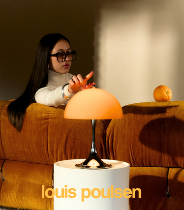 Vrouw met zonnebril raakt een oranje Louis Poulsen tafellamp uit de nieuwe Panthella collectie aan. De lamp staat op een wit bijzettafeltje voor een oranjekleurige bank, in warm, zacht licht.