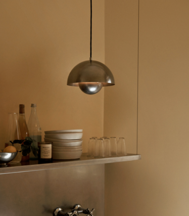 Hanglamp van gepolijst metaal boven een plank met keramische schalen en glazen, voor een zandkleurige muur.