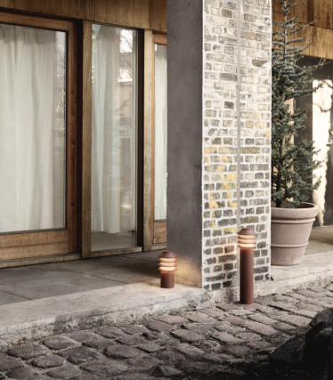 Buitenopstelling met twee moderne cilindervormige padverlichtingen naast een stenen voetpad en een kolom van baksteen en beton. Daarachter staan houten glazen deuren met lichtgordijnen en een grote groenblijvende plant in een pot. De warme gloed van de lampen zorgt voor een gezellige en uitnodigende sfeer.